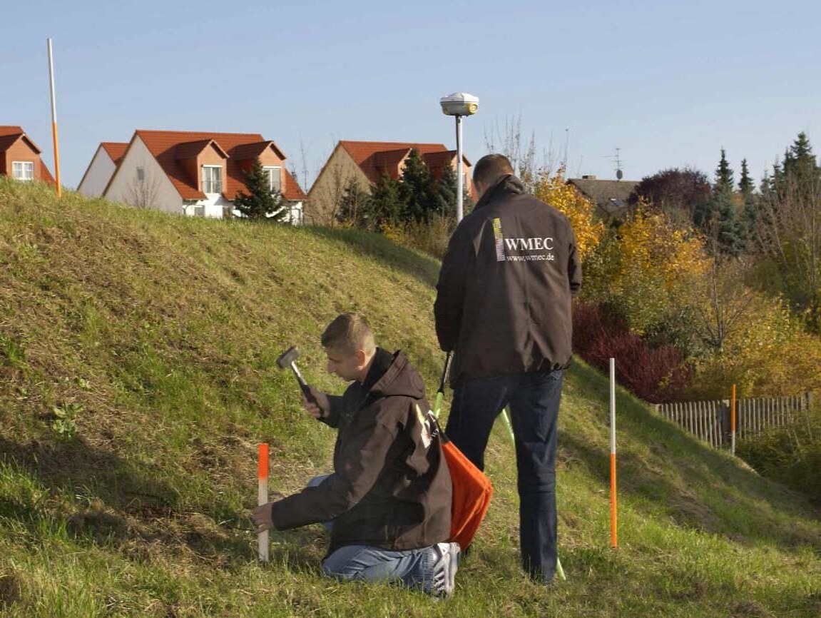 Zwei WMEC-Mitarbeiter bei der Absteckung eines Baugebiets mit GNSS-Empfänger und Holzpflöcken
